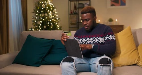 An Attractive Guy of African Appearance Sits on Couch Holding Tablet and Bank Card and Ordering