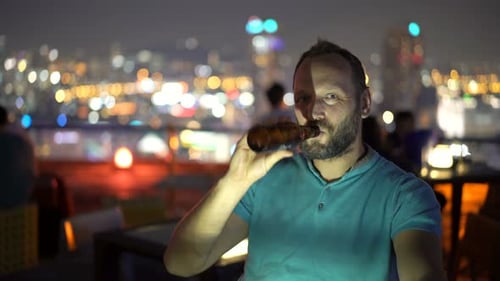 Young man enjoying refreshing beer at rooftop bar in city at night