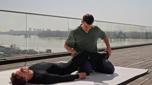 Physiotherapist in Green Scrubs Stretching Patient's Leg on a Sunny Rooftop with River and City