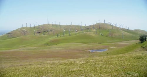 Wide shot of pond and green hill covered in windmills on a windy day.