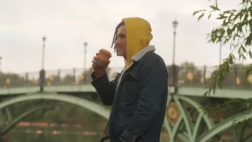 Young Man with Dreadlocks Holding Cup of Coffee Standing Near River Lake on Background of Big Bridge