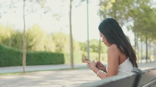 Young woman sitting on park bench typing message on mobile phone