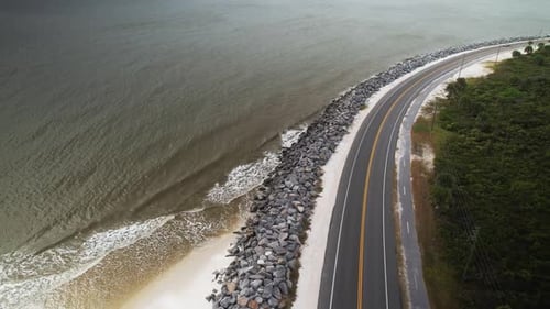 Aerial approach of vehicles travelling coastal road adjacent to waves crashing against rock barrier