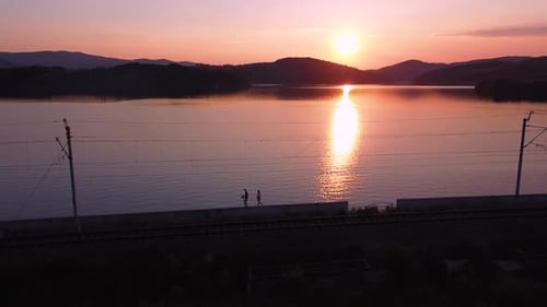 Aerial shot of walking couple, railway track, clear sky, sunset and huge orange lake