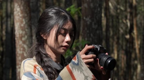Attractive young Asian woman is holding a camera, taking picture of the forest nature.