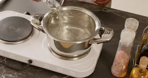 Man pouring water from measuring cup into pot on hot plate, reaching controls to heat water