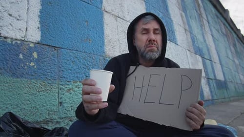 Old Homeless Poor Man Sitting on the Ground in the City with a Help Table While Asking for Money to