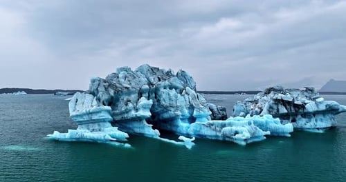 Vista de perto por drone de um iceberg azul flutuando em águas abertas e frias sob nuvens cinzentas, mostrando rugosidade