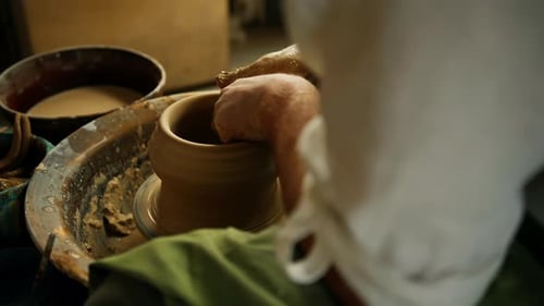 Close-up of potter's hands covered with clay making beautiful vase on throwing wheel in pottery