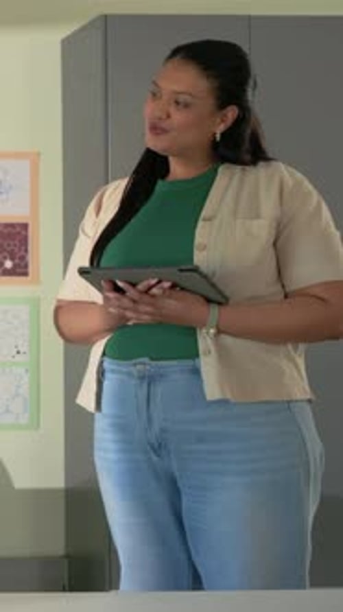 Vertical video: Responding to class, teacher in green top holding tablet with laptop in classroom