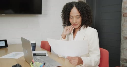 Smiling biracial businesswoman at desk with laptop holding document at office, in slow motion