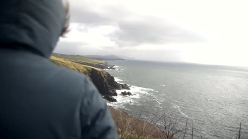 Person in green hooded jacket stands on edge and watches towards cliffs and wild sea in distance, wa