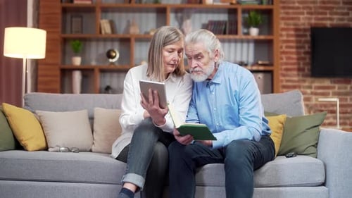 Elderly Couple Viewing Tablet and Book Together on Sofa