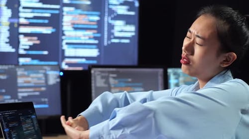 Asian Female Programmer Stretching While Writing Code By A Laptop Using Multiple Monitors