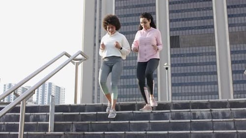 After pausing atop concrete stairs at plaza Diverse female friends jogging down near metal handrail