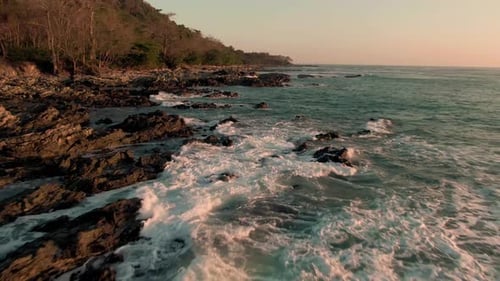 Drone parallax flight above breaking swell on rocky Costa Rica beach in sunset