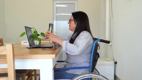 Una mujer en silla de ruedas se pone los auriculares y comienza a trabajar con el portátil en casa. - plano panorámico