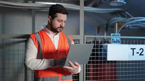 Electrical Engineer Working on Laptop Near Electrical Switchboard in Industrial Environment