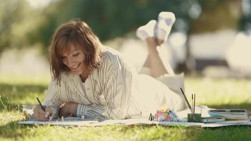 An Adult Woman Lying on the Blanket in the Blooming Park and Drawing in the Sketchbook