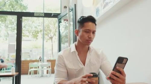 Young businessman sits at table with mobile phone and drinks coffee in cafe