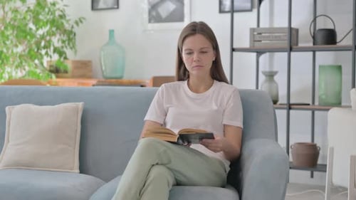 Young Woman Reading Book while Relaxing on Sofa at Home