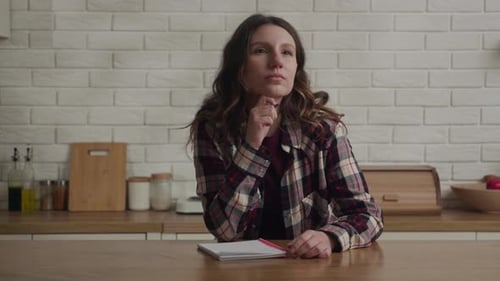 A Young Woman in a Checkered Shirt Sits at a Kitchen Table Writing Notes in a Notebook