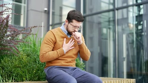 Sick businessman coughing sitting on a bench on the street near office building. Upset bearded man