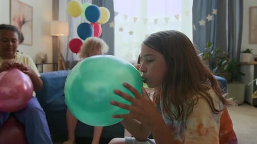 Long-haired Boy Blowing up Green Balloon while Preparing for Birthday Party