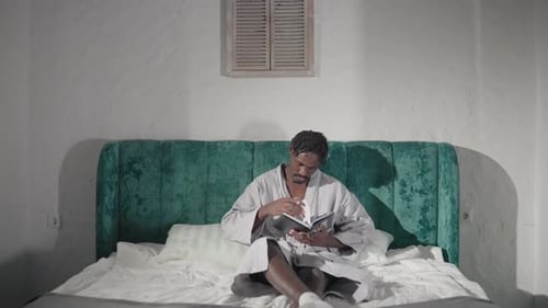 Man Reading Book in Cozy Robe on Bed with Green Velvet Headboard in Peaceful Bedroom