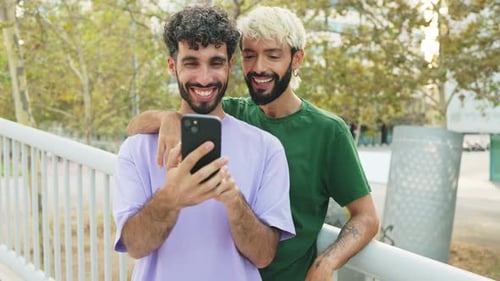 Cheerful gay couple using mobile phone in a park in daylight.