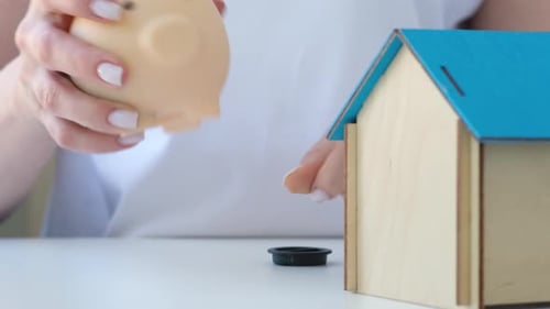 Woman Open and Shaking Coins Out of Piggy Bank Next to House Saving to Buy Home