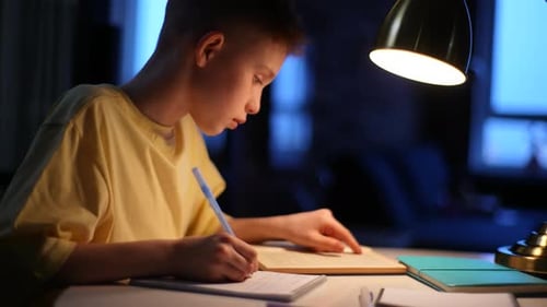Closeup Side View of Redhead School Boy Pupil Studying at Home Sitting at Table Under Light of Lamp