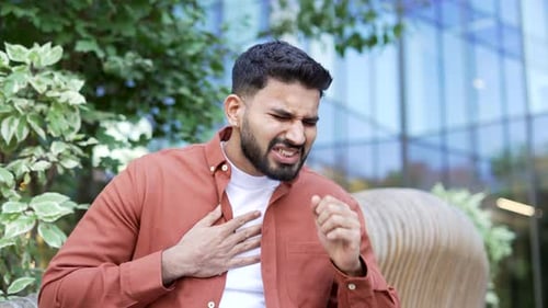 A sick businessman coughing sitting on a bench on the street near office building. Upset bearded man