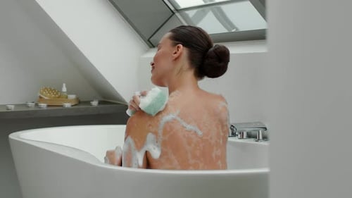 Woman in bathtub scrubs her back with sponge