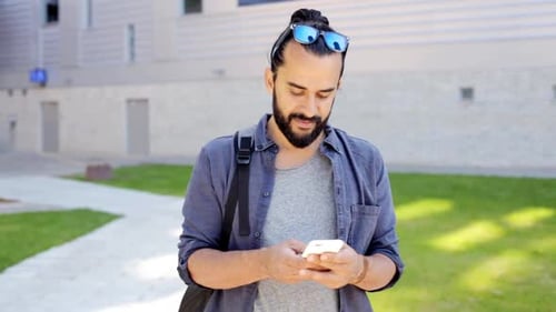 Smiling man with backpack enjoys summer vacation while texting on smartphone in city