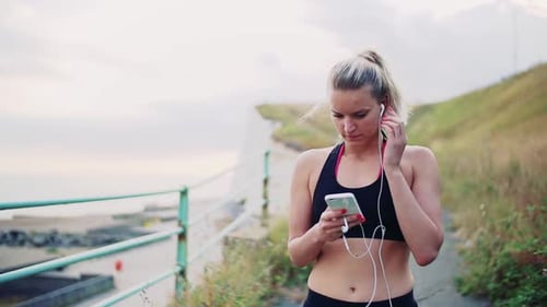 Young Sporty Woman Runner with Smartphone and Earphones on the Beach Outside