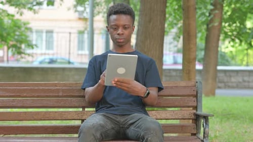 Casual African Man Using a Digital Tablet on a Park Bench