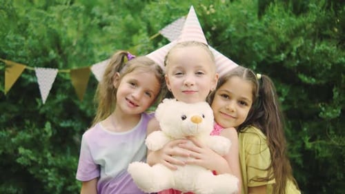 Três meninas usando chapéus de festa se divertindo ao ar livre, abraçando um ursinho de pelúcia e acenando para