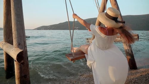 Slow motion footage of a young brunette woman in a flowing white dress gracefully swinging at sea