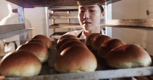 Fröhliche asiatische Bäckerin, die in der Bäckereiküche arbeitet und Backblech mit Brötchen in Zeitlupe hält
