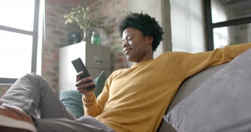 Happy african american man sitting on sofa using smartphone at home, slow motion