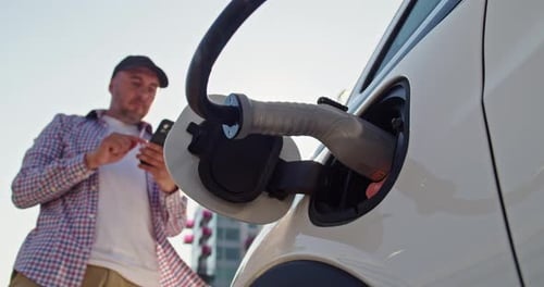 Man with Smartphone Waits While His Electric Car Charging at a Charging Station