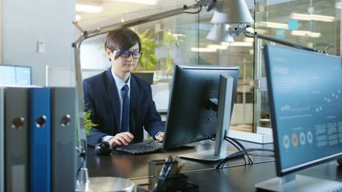 In the Office East Asian Businessman works on a Desktop Personal Computer. His Colleague's Computer