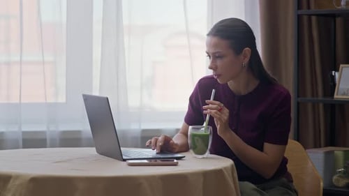 Young Woman Drinking Green Smoothie while Looking at Laptop Screen
