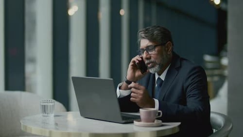 Mid-Aged Businessman Talking on Phone as Working on Laptop in Cafe