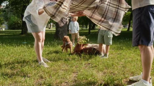 Parents Setting Up Blanket for Picnic in Park, Children Playing with Dog