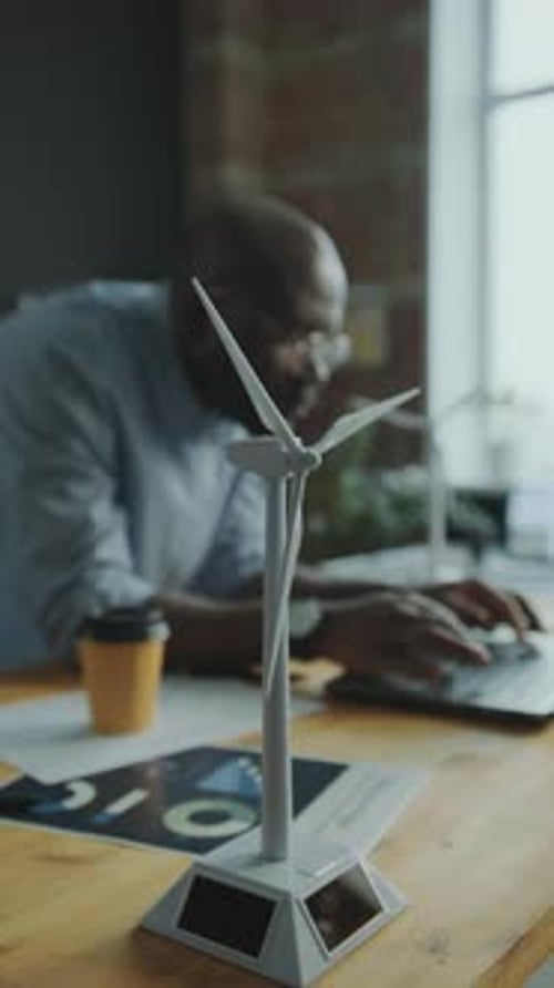Close-Up of Windmill Turbine on Office Desk, Engineer Using Laptop Behind