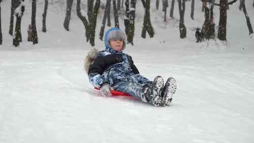 Slow motion footage of a boy gleefully gliding down a snow-covered hill on his plastic sled
