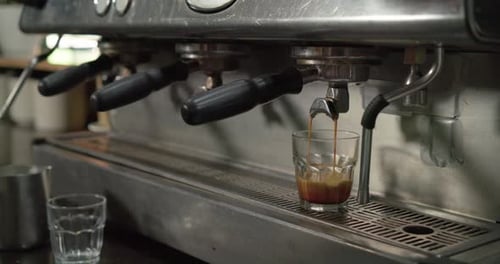 Machine spout is starting to release espresso into glass on drip tray, extracting crema for serving