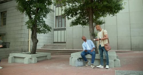 Two men sitting on bench in plaza, blue-shirt laughing at phone while tan-tee coming showing snack
